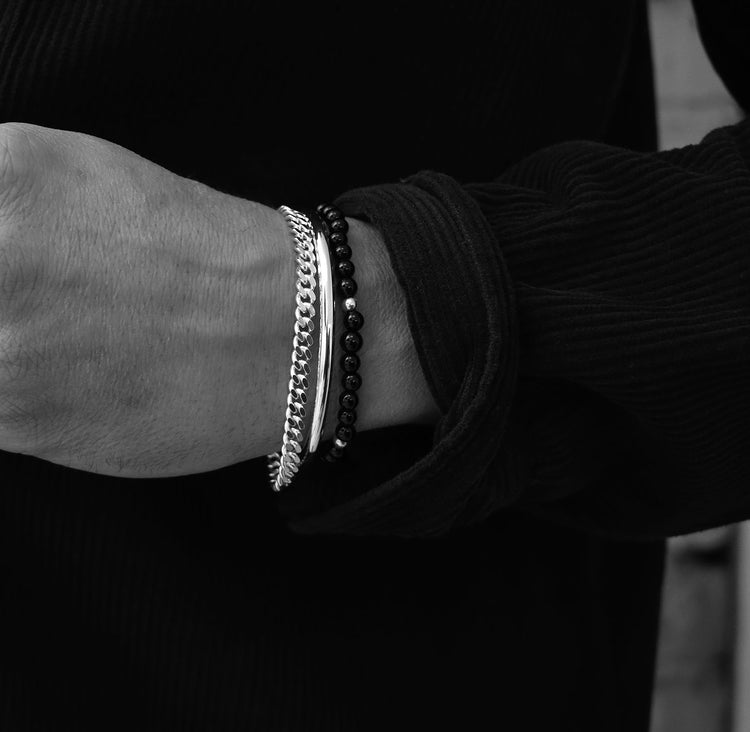 Man som bär ett silverarmband med kedja, ett cuff armband i silver och ett armband med svarta stenar.
