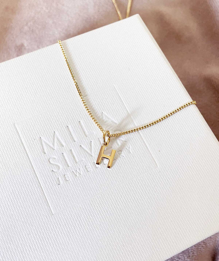 Bokstavsberlock i guld i guldkedja placerad pÄ present ask med Mila Silvers logotyp.