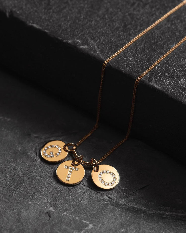 tre guldberlocker med diamantbokstav i guldkedja placerad på ett svart stenblock.