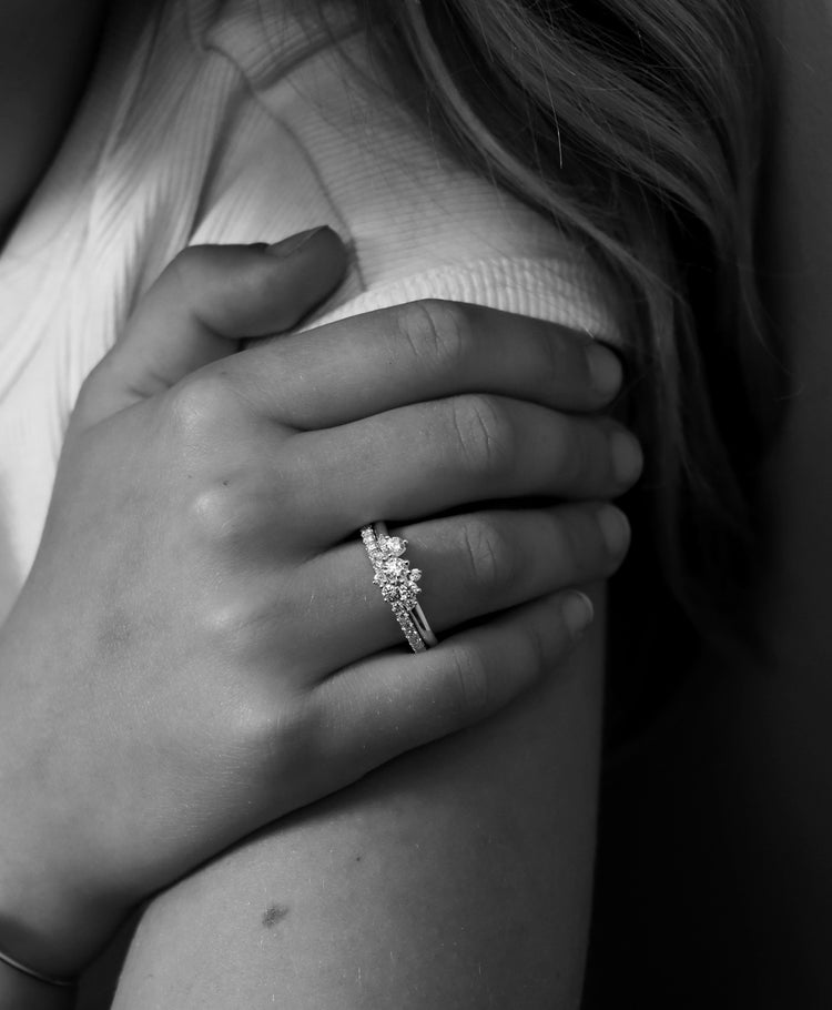 Två diamantringar i kombination. En alliansring och en ring med flera diamanter i ett kluster. Bärs på vänster ringfinger av en ung kvinna med långt hår.