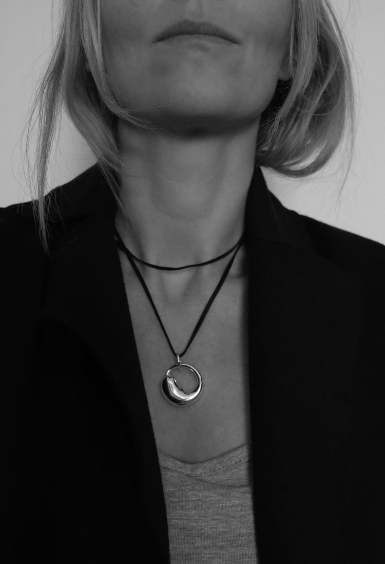 Kvinna som bär ett halsband med runt hängsmycke i silver som hänger i svart läderband, virat två varv runt halsen.