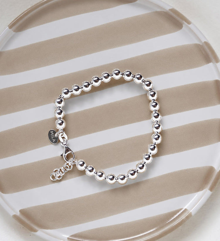 Armband med kulor i silver som har ställbar längd, placerat på ett randigt fat. 