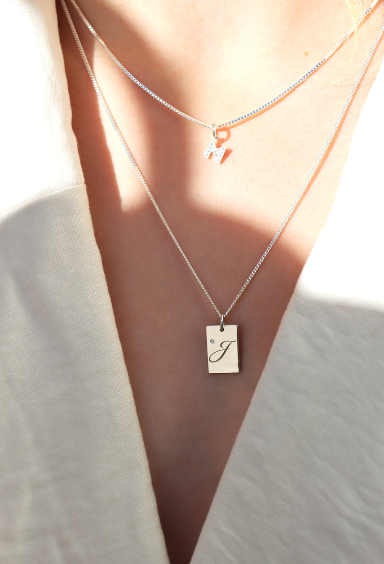 Tjej som bär bokstavshalsband. En diamantbokstav i kort kedja och en berlock med rektangulär bricka och graverad bokstav + diamant.