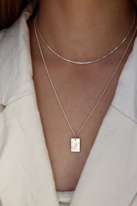 tjej som bär ett bokstavhalsband i silver kombinerat med en tunn kort kedja. 