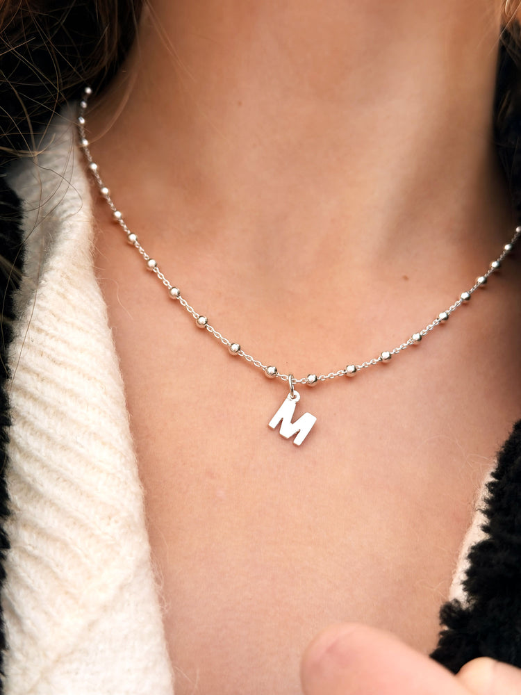 Tjej som bär ett bokstavshalsband i silver. Kedjan har små kulor med berlock - bokstav M.