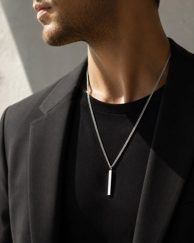 Man som bär ett längre silverhalsband med stav i kedja. han har svart kostym med svart t-shirt under