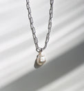 Halsband som hänger mot en solig vägg med en silverkedja som har ovala länkar och en stor oregelbunden barockpärla. 