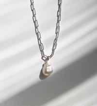 Halsband som hänger mot en solig vägg med en silverkedja som har ovala länkar och en stor oregelbunden barockpärla. 