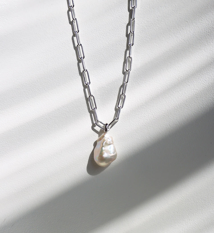 Halsband som hänger mot en solig vägg med en silverkedja som har ovala länkar och en stor oregelbunden barockpärla. 