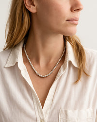 Kvinna som bär ett kort halsband med ett band av silverkulor. Hon har vit/beige linneskjorta, uppknäppt och utsläppt hår. 